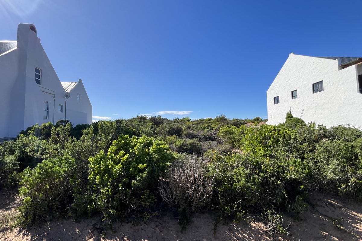 A view of a vacant residential plot situated between two white-walled, gabled architectural structures with natural coastal vegetation.