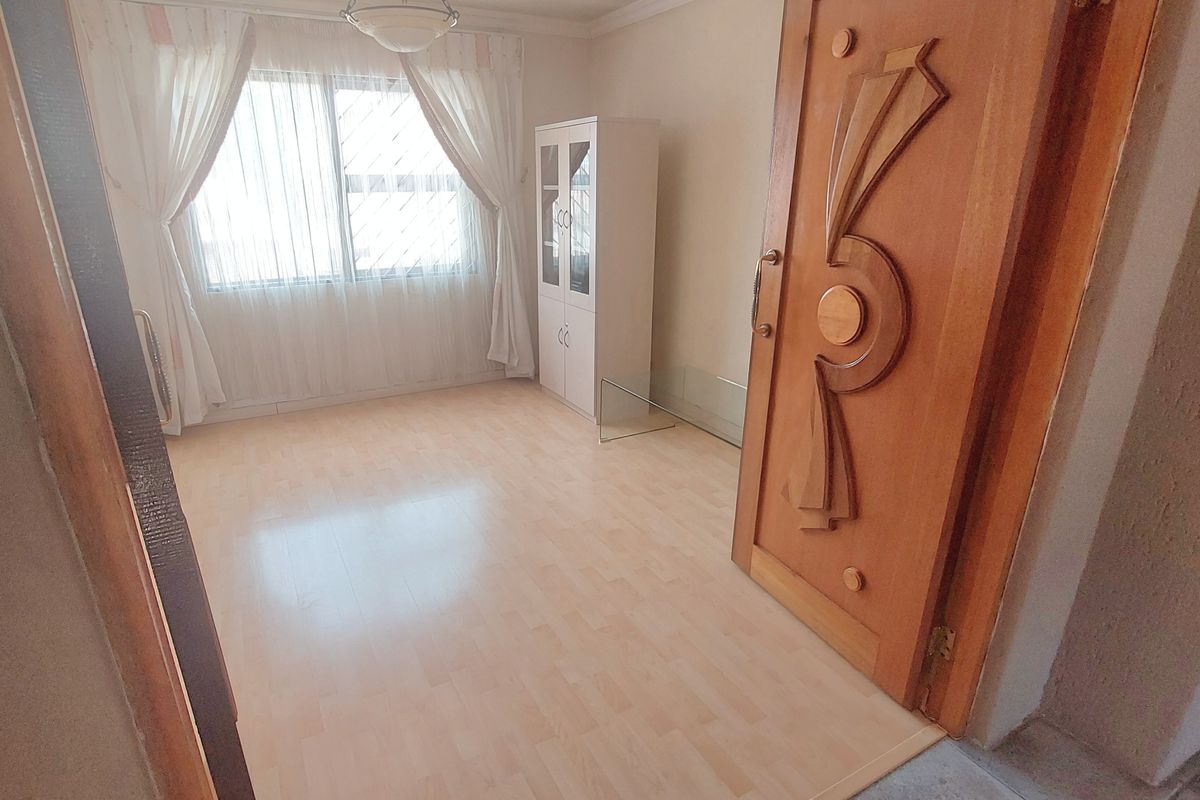 A bright, empty bedroom featuring light-toned laminate flooring, a decorative wooden door, and a classic ceiling pendant light fixture.