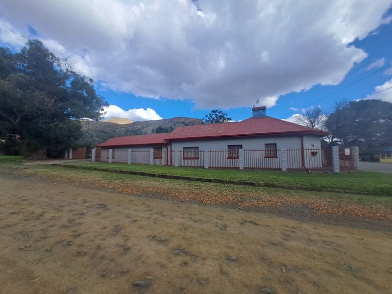 A single-story house featuring a red metal roof, perimeter fencing, and security bars on the windows, set against a mountainous backdrop.