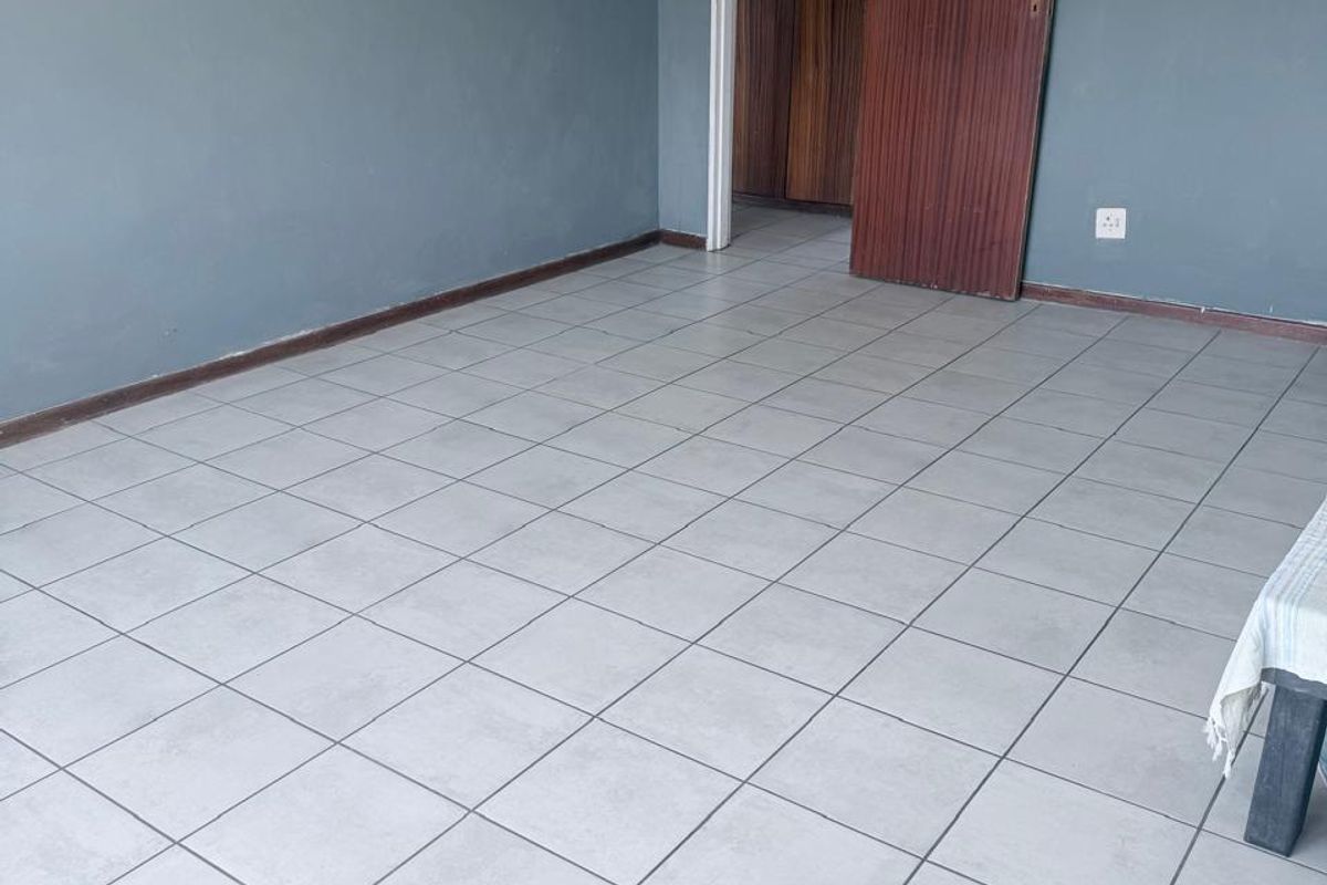 An empty living area featuring neutral tiled flooring and blue-toned walls, leading to a room with built-in wooden cupboards.