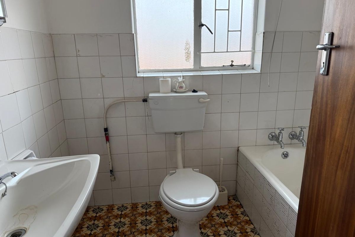 A dated bathroom featuring a porcelain bathtub, pedestal basin, and close-coupled toilet, finished with original patterned floor tiles and white wall tiling.