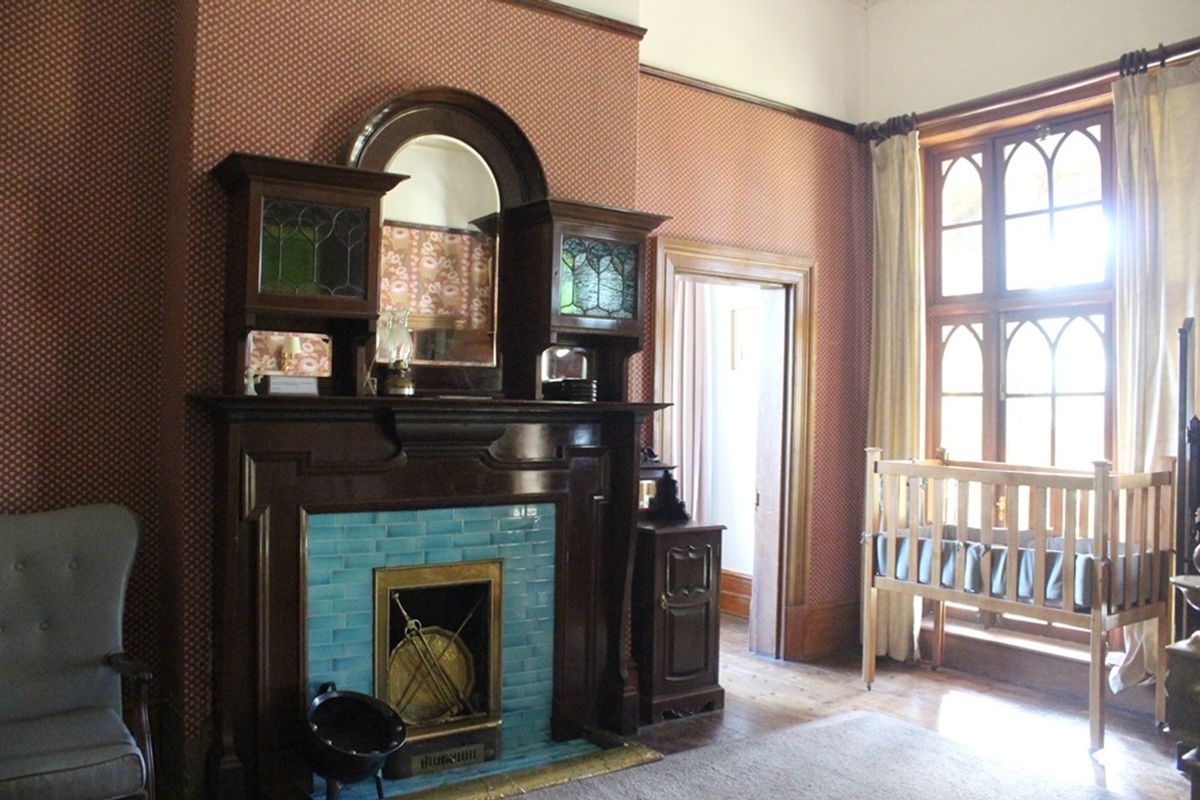 A character-filled bedroom featuring a prominent period fireplace with decorative blue tiling, timber mantelpiece, and large arched wooden window frames.