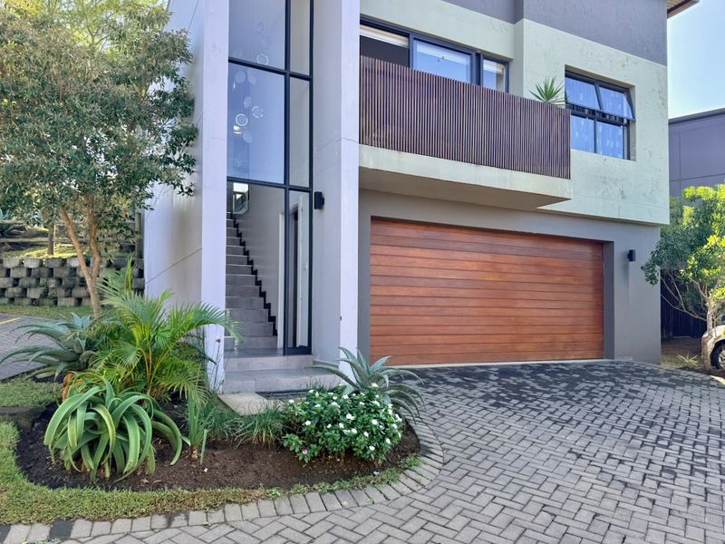 Modern townhouse exterior featuring a timber garage door, a private balcony with timber balustrade, and a striking full-height glass entrance with staircase.