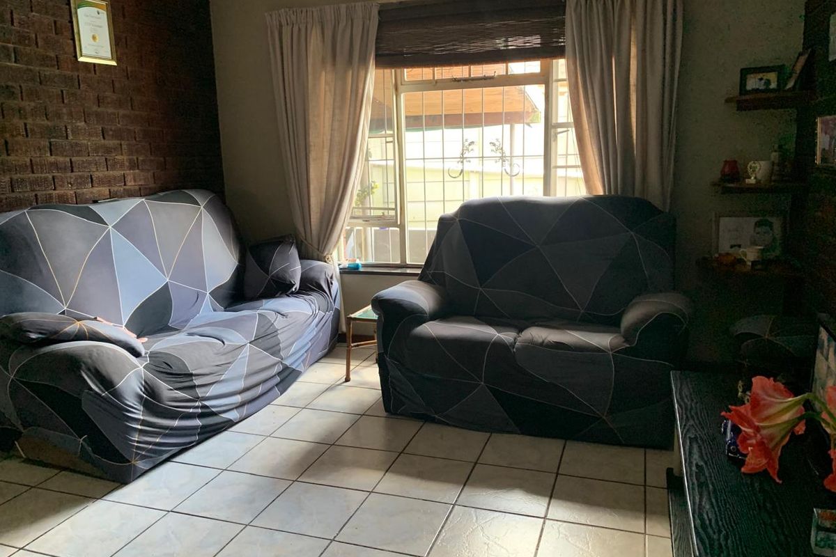 A living room featuring tiled flooring, an exposed brick feature wall, and a window fitted with security bars.