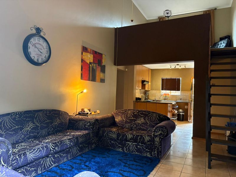 Open plan living area featuring a vaulted ceiling, mezzanine level with staircase, and an adjoining kitchen with built-in cabinetry and tiled flooring.