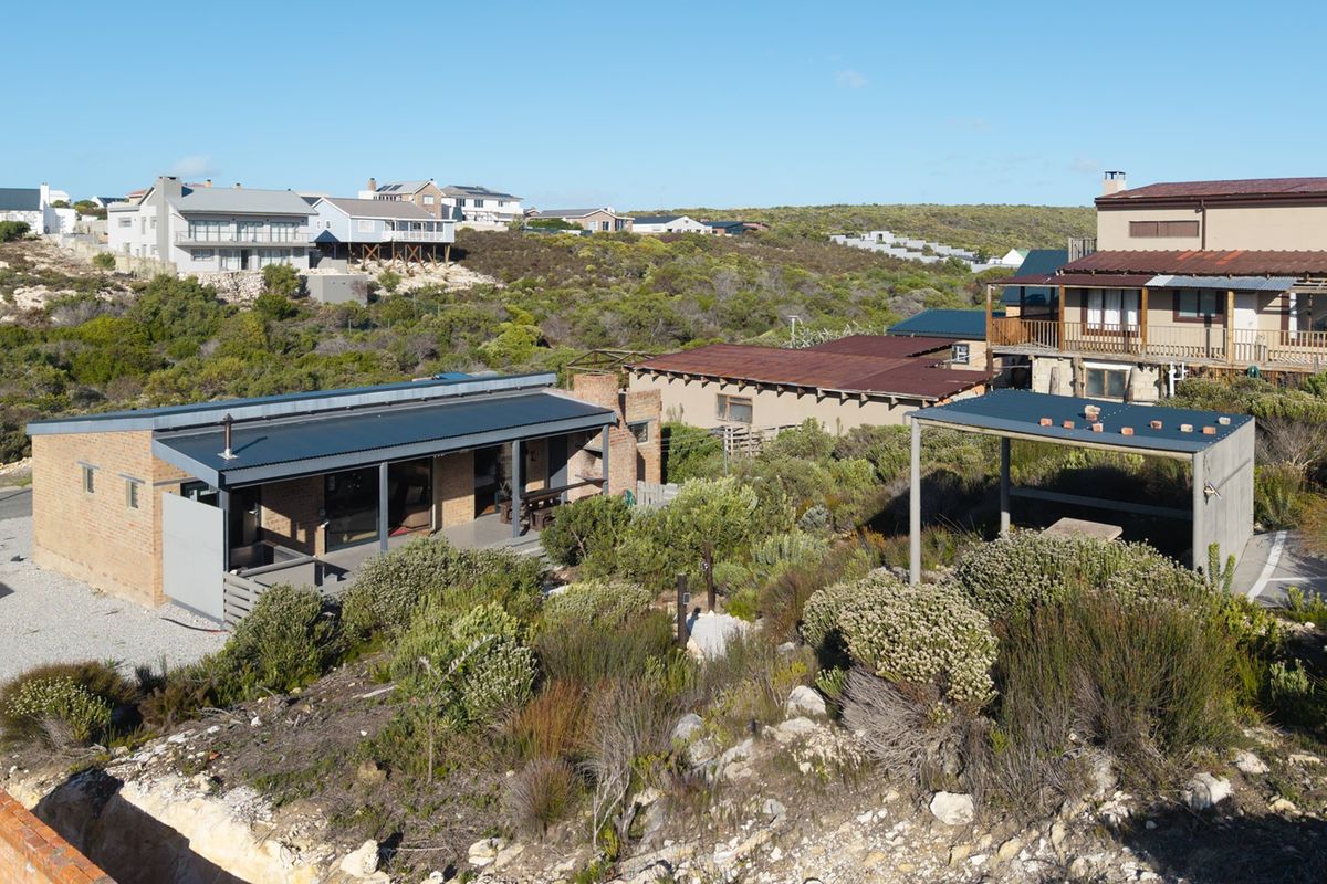 Aerial view of a coastal residential property featuring a brick house with a corrugated metal roof, covered patio, and an outdoor pergola structure.