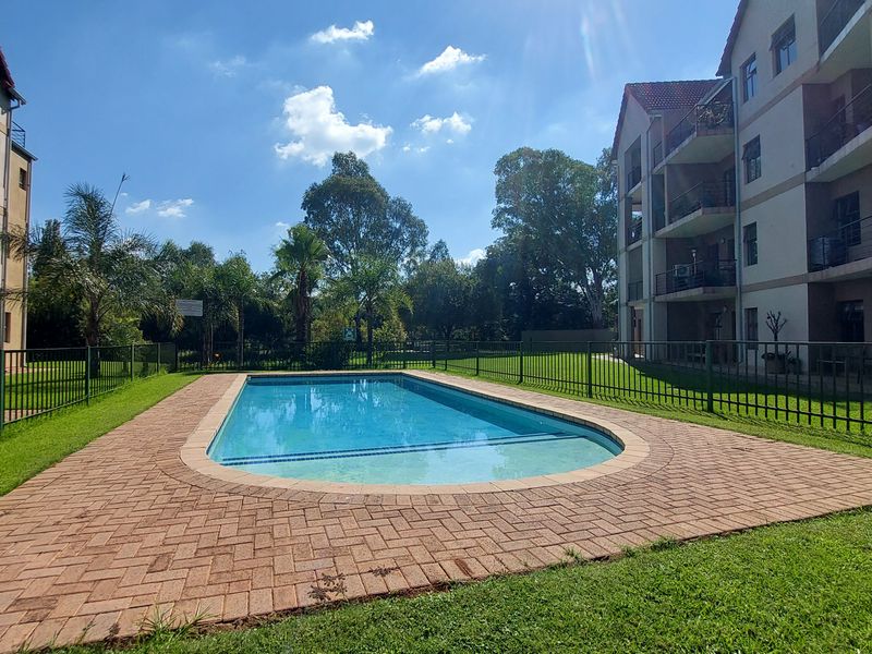 A well-maintained communal swimming pool area featuring paved surrounds, manicured lawns, and secure metal perimeter fencing within an apartment complex.