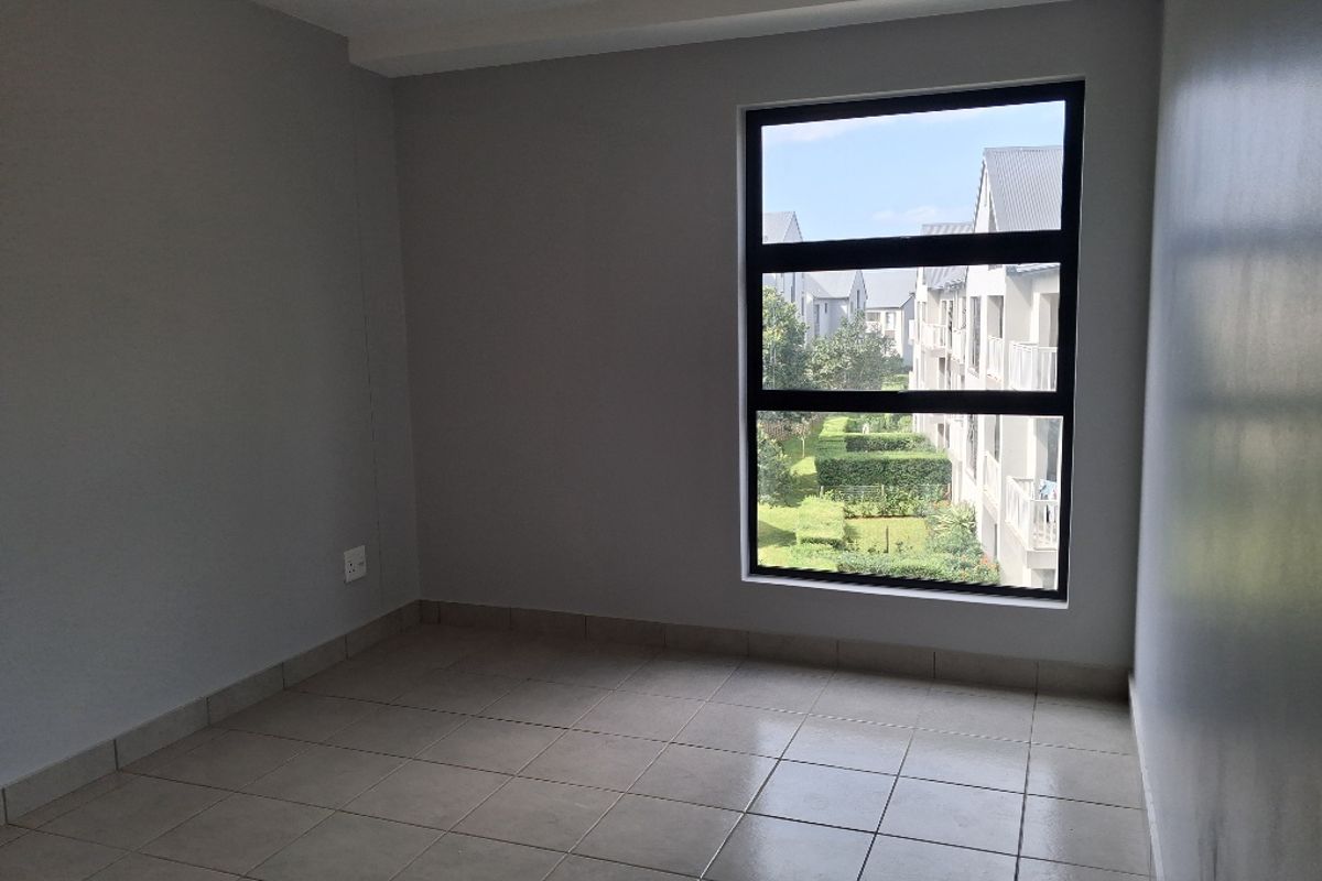 A bright, empty bedroom featuring neutral tiled flooring, a large window with a view of the complex grounds, and a clean, modern finish.