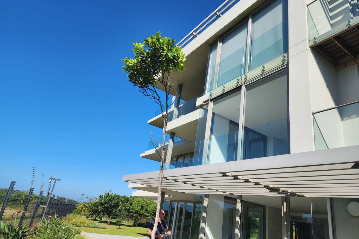 Modern apartment building exterior featuring expansive glass sliding doors, sleek glass balustrades, and a covered patio area overlooking the landscaped grounds.