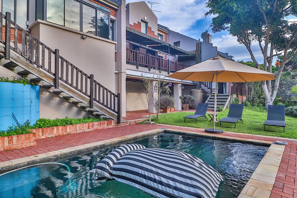 Rear exterior view featuring a swimming pool, brick paved patio area, landscaped garden, and a multi-level building facade with timber balconies and stairs.