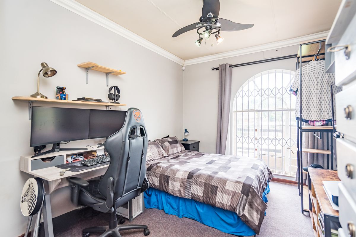 A functional bedroom featuring an arched window with security gate, ceiling fan, and carpeted flooring, currently utilized as a combined sleeping and workspace area.
