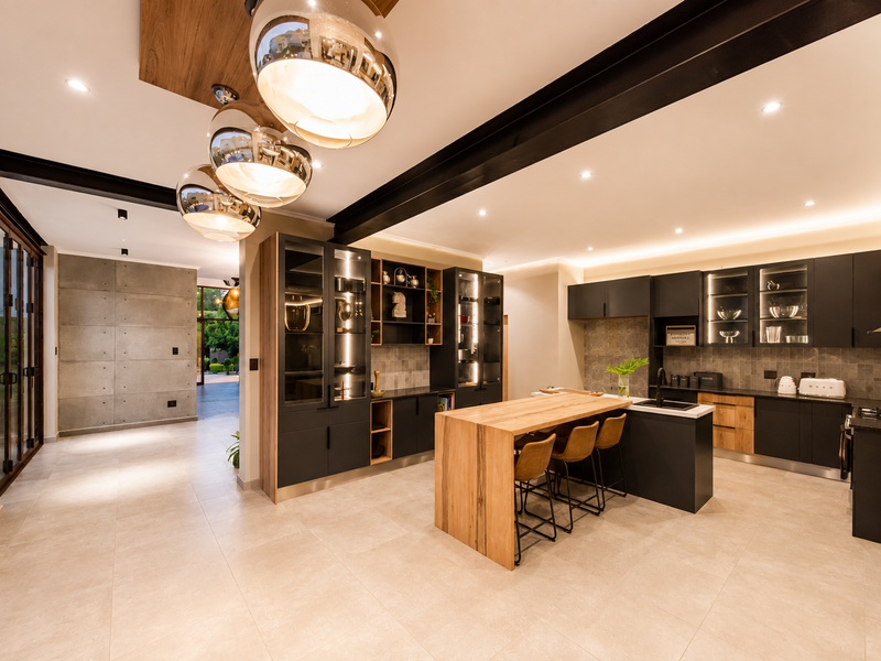 Modern open-plan kitchen featuring dark cabinetry, a timber breakfast bar, integrated lighting, and a striking concrete feature wall with high-end finishes.