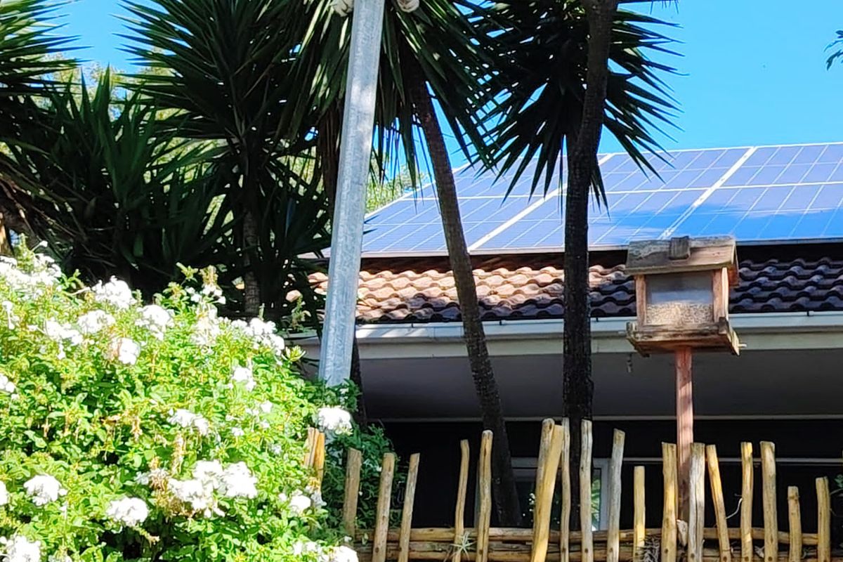 A view of the rear roofline featuring installed solar panels, a security camera mounted on a pole, and a rustic wooden fence.