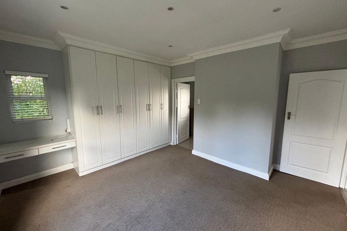 A spacious bedroom featuring neutral grey walls, carpeted flooring, extensive built-in cabinetry with an integrated desk, and recessed ceiling lighting.
