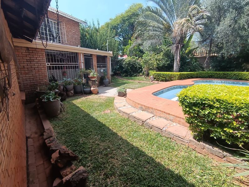 A backyard area featuring a swimming pool with brick surround, established garden landscaping, and a brick-walled house exterior with security bars.