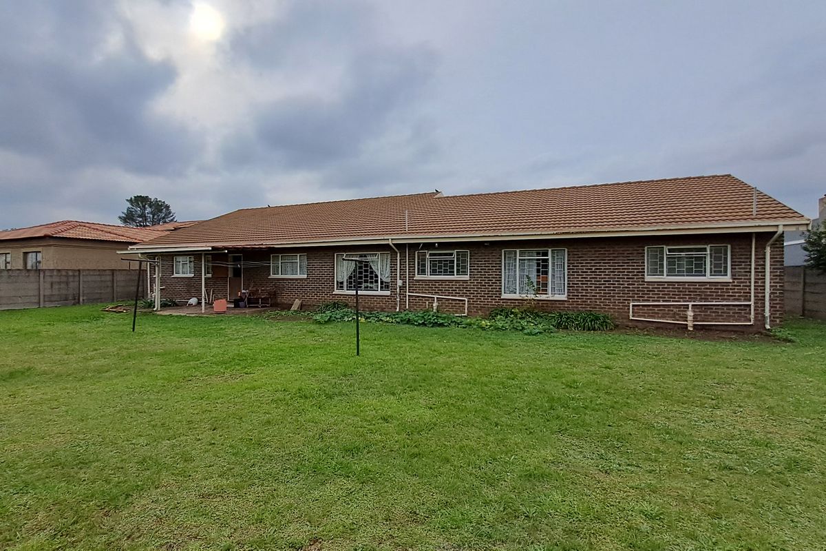 Rear view of a single-story brick house featuring a tiled roof, security bars on windows, and a covered patio area overlooking a spacious lawn.