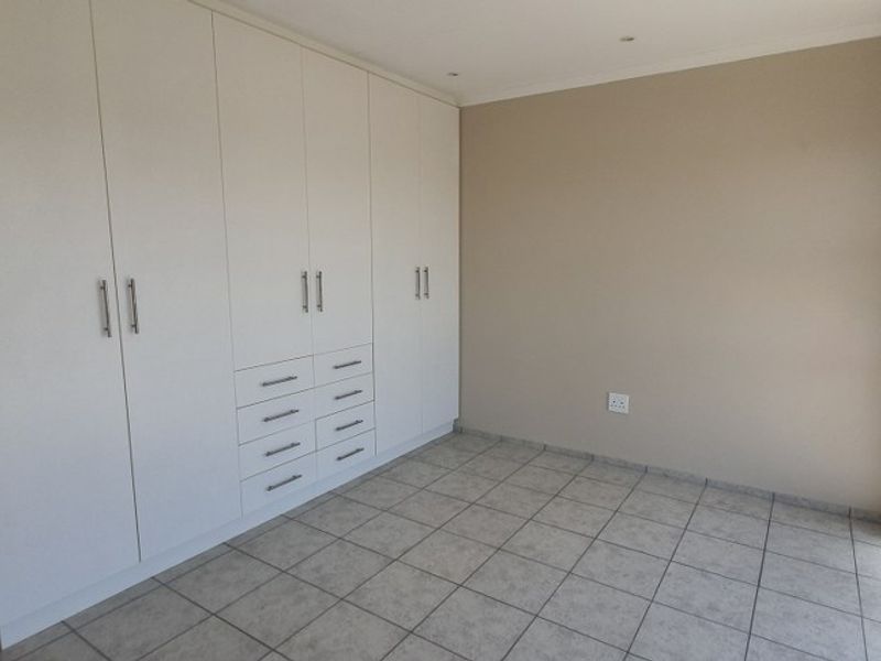 A bright, empty bedroom featuring neutral tiled flooring, extensive built-in cupboard storage, and a sliding door providing access to an outdoor balcony.