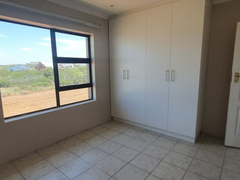 A bright, empty bedroom featuring neutral tiled flooring, built-in cupboards, and a large window with views of the surrounding landscape.