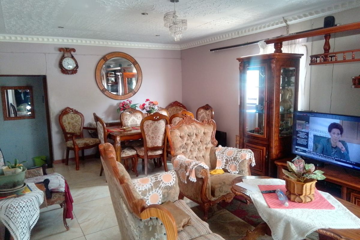 A living room featuring tiled flooring, ornate ceiling cornicing, and a central crystal chandelier, showing signs of wear and dated finishes.