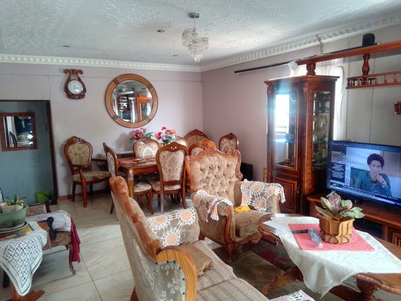 A living room featuring tiled flooring, ornate ceiling cornicing, and a central crystal chandelier, showing signs of wear and dated finishes.