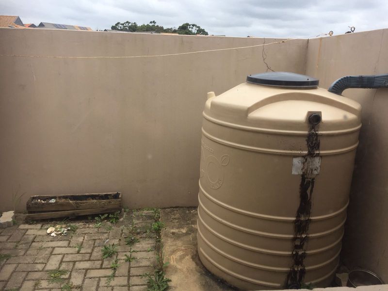 Utility area featuring a water storage tank connected to a downpipe, set against a boundary wall with paved flooring.