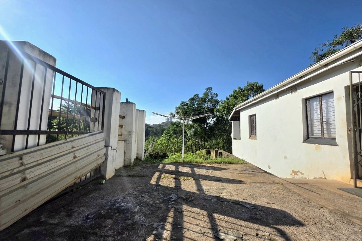 Rear exterior area featuring concrete paving, security fencing, and access to the side of the house with visible wear to finishes.
