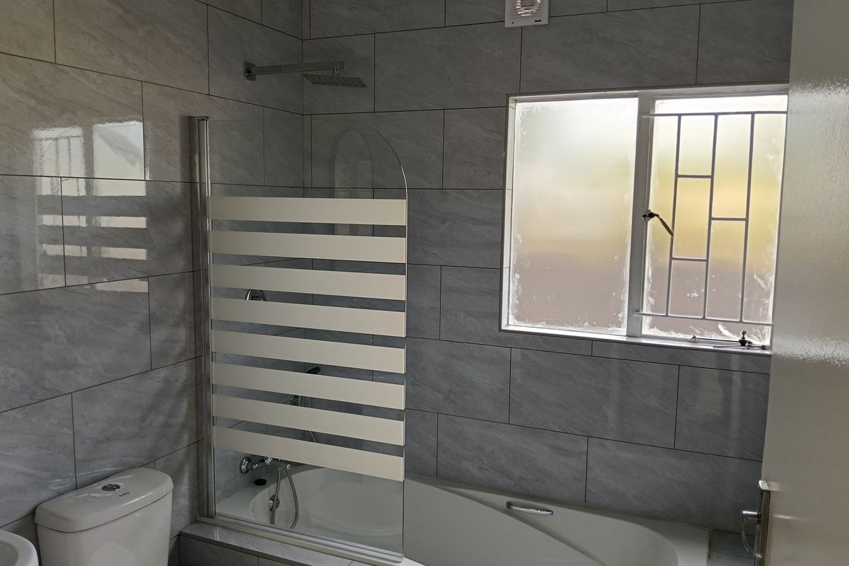 Modern bathroom featuring grey wall tiles, a bathtub with a glass shower screen, overhead shower fixture, and a window with security bars.