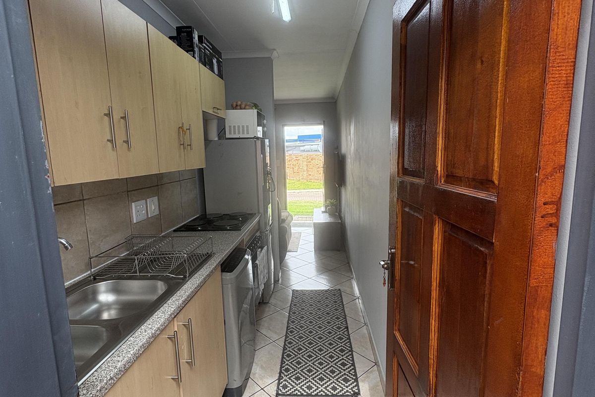 A narrow galley-style kitchen featuring light wood cabinetry, a stainless steel sink, and tiled flooring, leading directly to an exterior doorway.