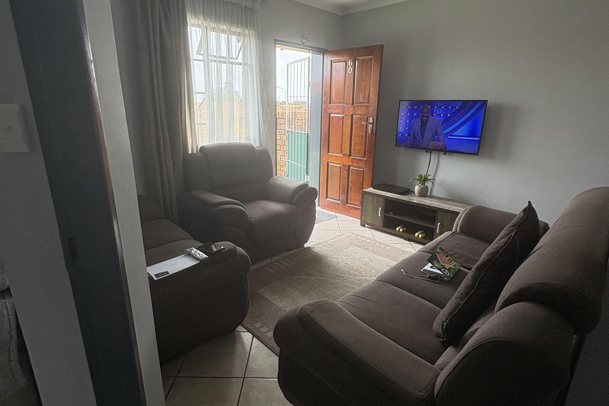 A compact living room featuring tiled flooring, a wooden entrance door, and a window with curtains, providing a functional and accessible living space.