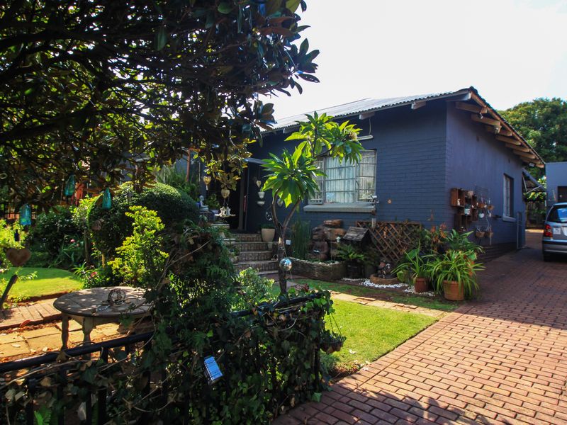 Front view of a painted brick house featuring a paved driveway, established garden, and metal roof.