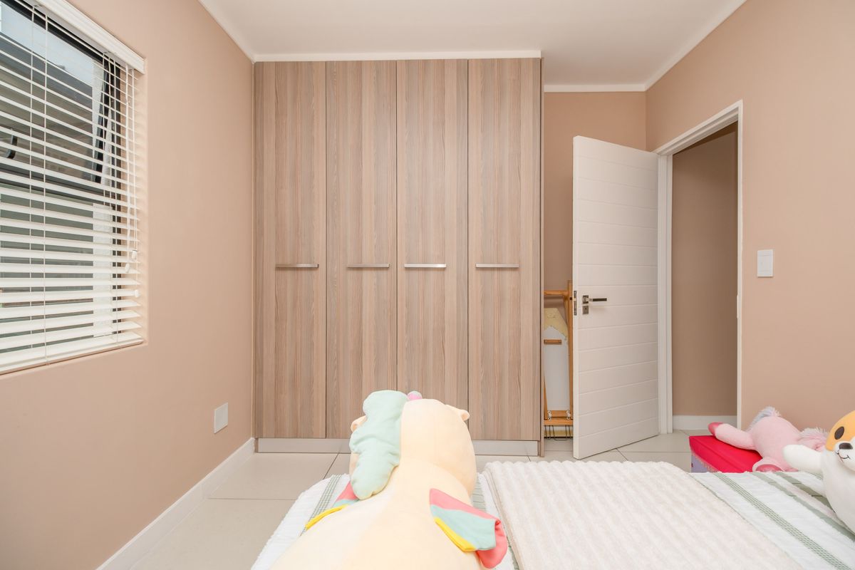 A well-maintained bedroom featuring light wood-grain built-in cupboards, tiled flooring, and a window fitted with horizontal blinds.