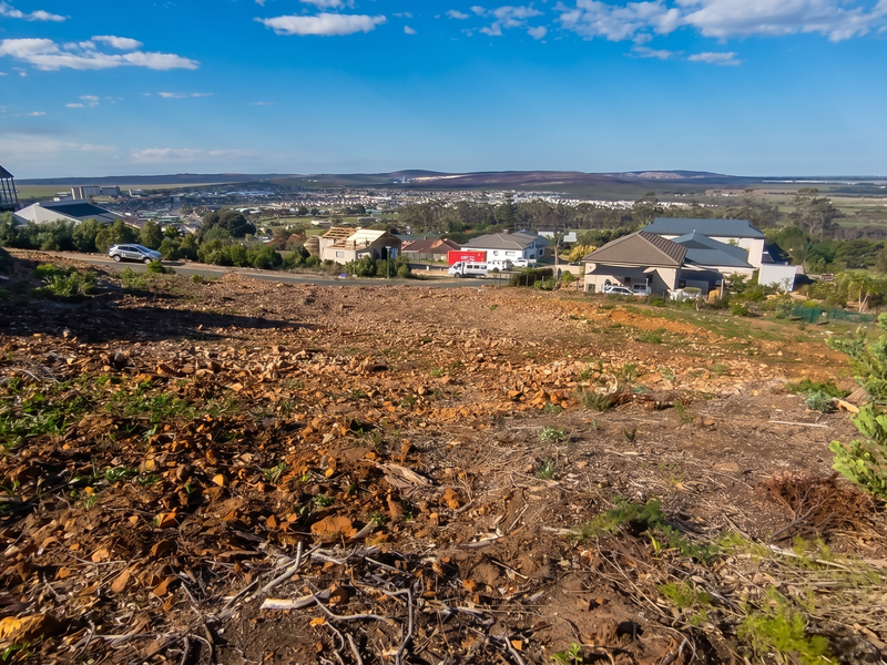 A wide-angle view overlooking a sloped, undeveloped residential plot with surrounding neighborhood and distant landscape vistas.