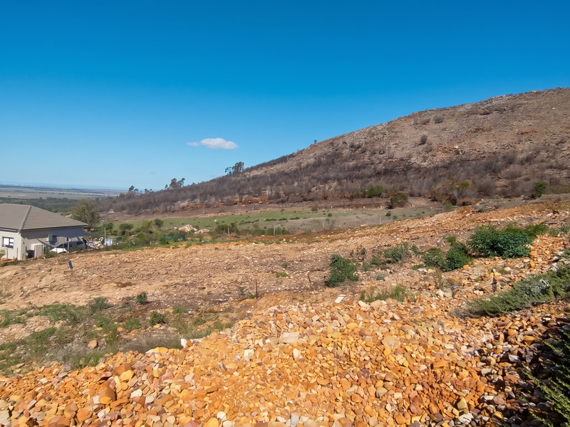A wide-angle view of a sloped, undeveloped residential plot featuring rocky terrain with a mountain backdrop and neighboring properties.