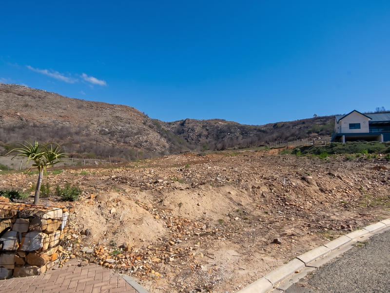 A vacant residential plot featuring a gabion retaining wall, paved driveway access, and scenic mountain views in a quiet estate setting.