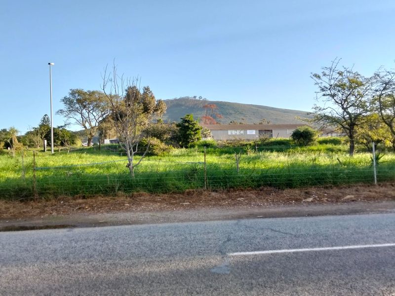 A view of a vacant residential plot from the street, featuring grassy terrain, perimeter wire fencing, and a mountain backdrop.