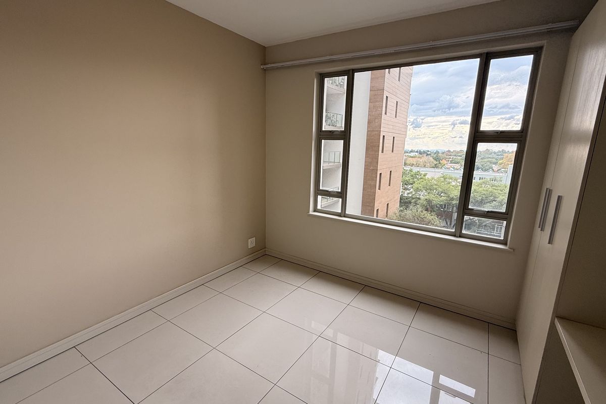 A bright, empty bedroom featuring large windows with exterior views, neutral tiled flooring, and built-in storage cupboards.
