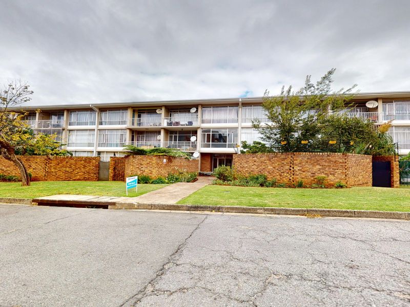Front exterior view of a multi-story apartment complex featuring brick boundary walls, private balconies, and a paved entrance walkway.