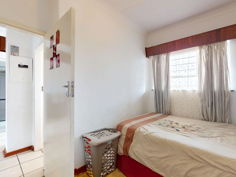 A compact bedroom featuring a window with security bars and a wooden pelmet, adjacent to a tiled hallway with an electrical distribution board.