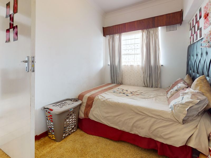 A compact bedroom featuring carpeted flooring, a window with security bars, and a wooden pelmet, offering a functional space for rest.