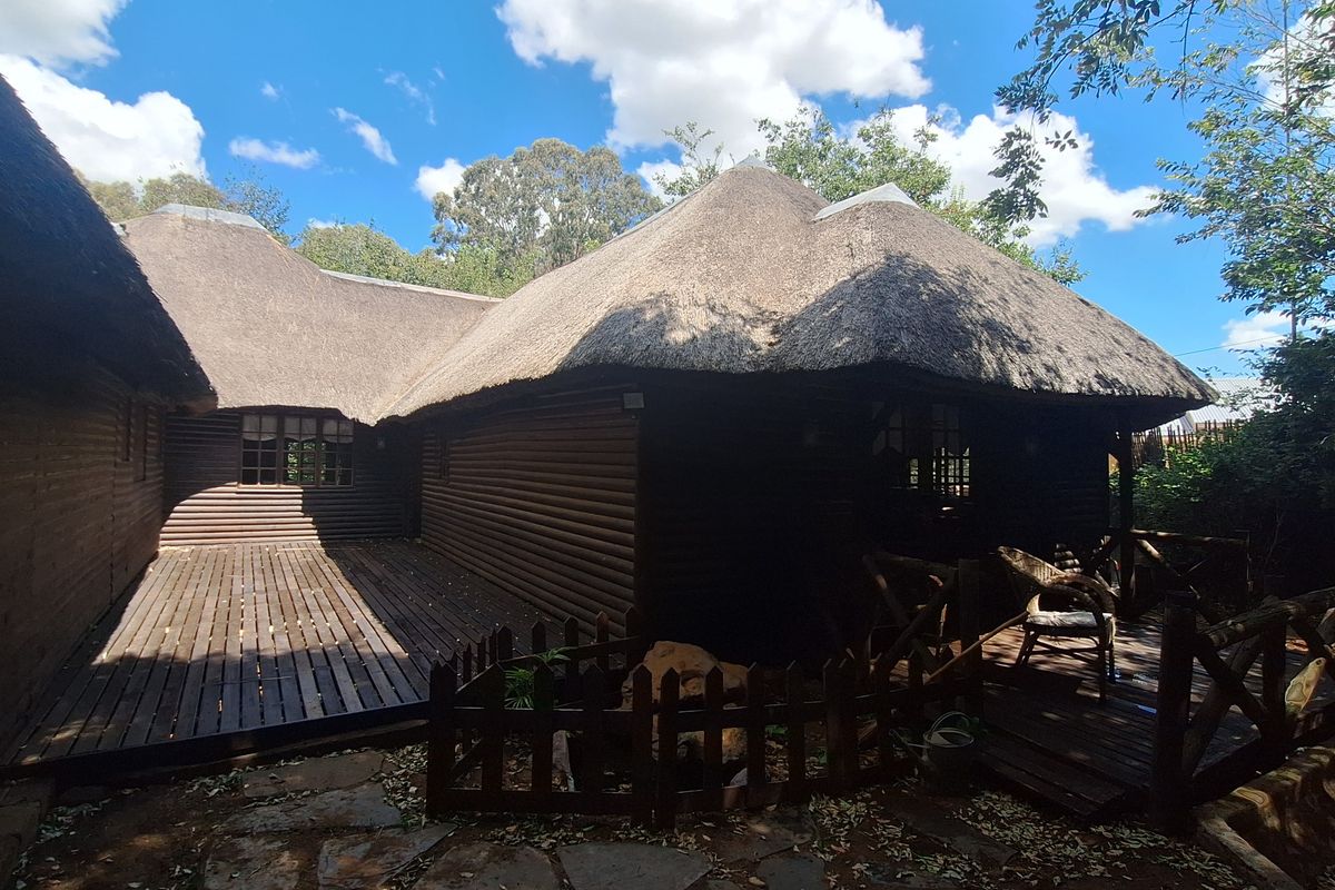 Rear view of a rustic log cabin featuring a traditional thatch roof, extensive timber decking, and a stone-paved garden area.
