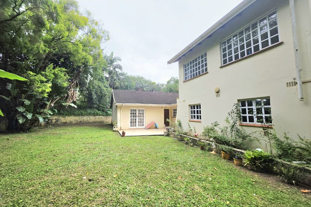 Rear view of the property featuring a spacious lawn, a paved patio area, and a multi-story building with grid-pattern windows and plastered exterior walls.