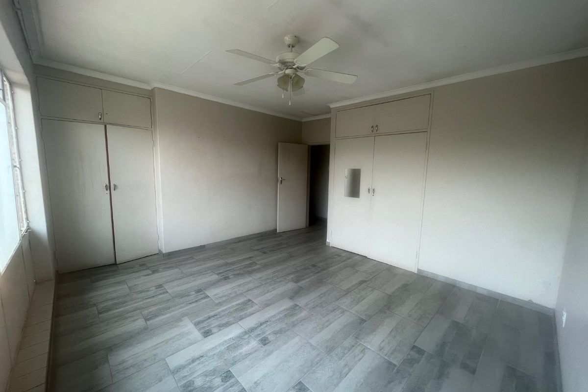 A spacious bedroom featuring neutral tiled flooring, built-in cupboards with overhead storage, and a ceiling fan.