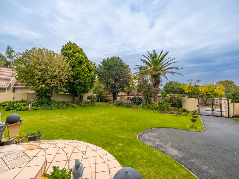 A well-maintained garden featuring a manicured lawn, mature trees, a paved driveway, and a tiled patio area enclosed by perimeter walls.