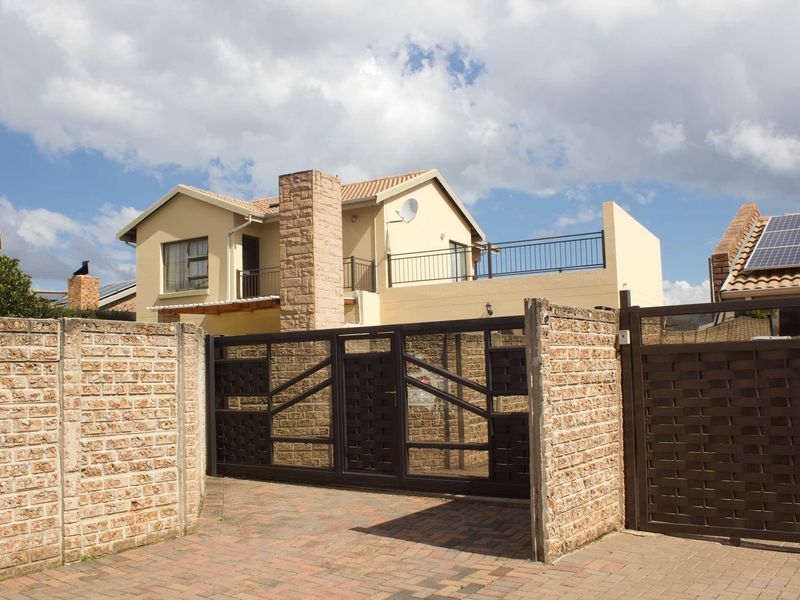 Front view of a double-story house featuring a paved driveway, secure metal gate, brick boundary wall, and an upper-level balcony.