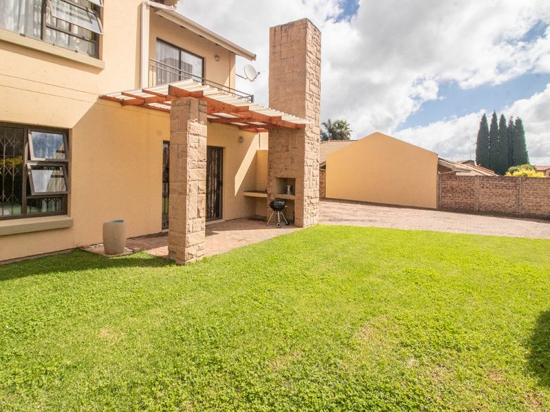 Rear exterior view featuring a paved patio area with a built-in stone braai, pergola, and a well-maintained garden lawn.