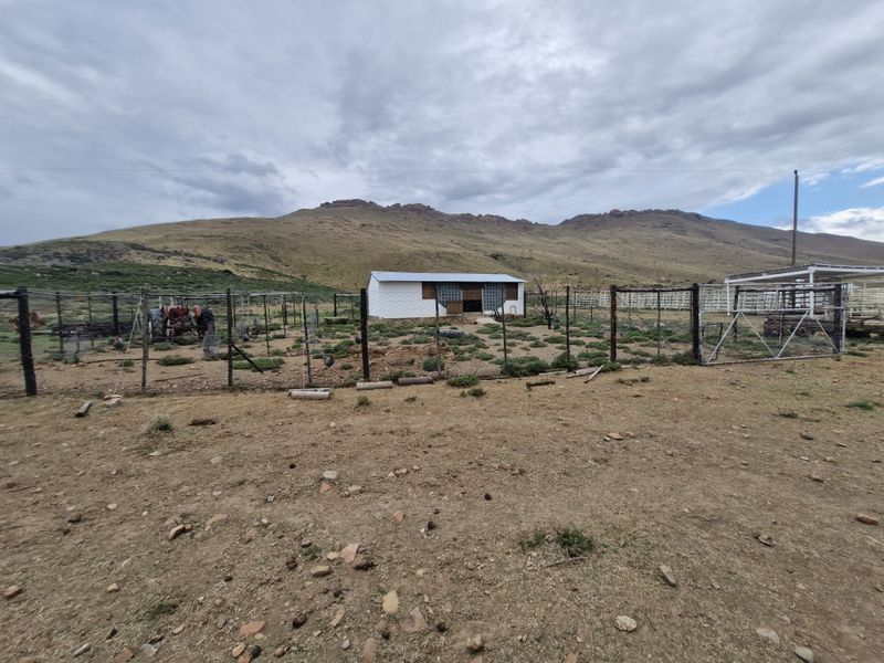 A rural farm yard featuring wire fencing, a metal gate, and a small outbuilding set against a mountain landscape.