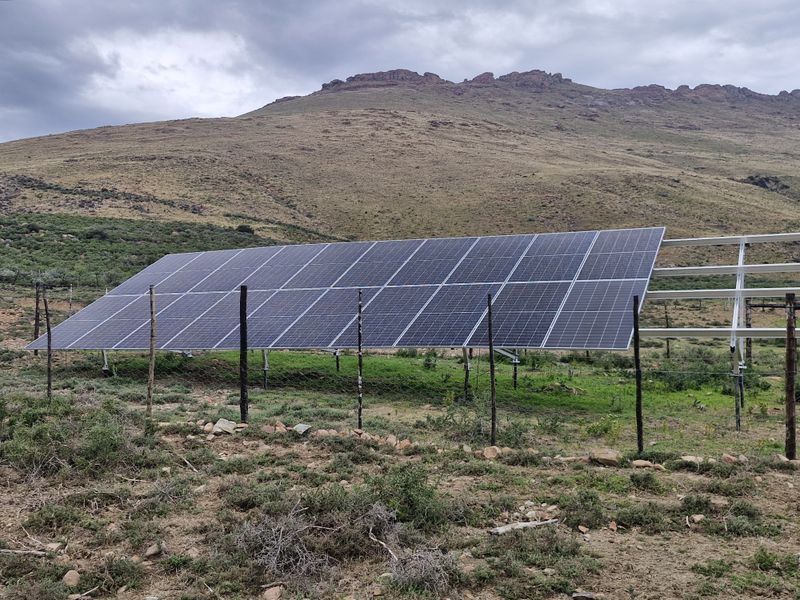 A ground-mounted solar panel array installed on a metal support structure, situated in a rural landscape with mountain views.