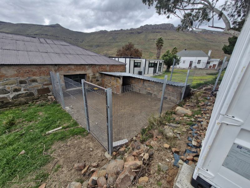 An outdoor utility area featuring stone masonry walls, corrugated metal roofing, and wire mesh fencing, set against a rural landscape.