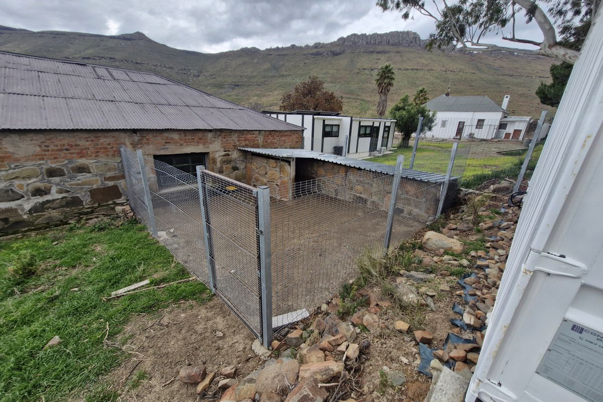 An outdoor utility area featuring stone masonry walls, corrugated metal roofing, and wire mesh fencing, set against a rural landscape.