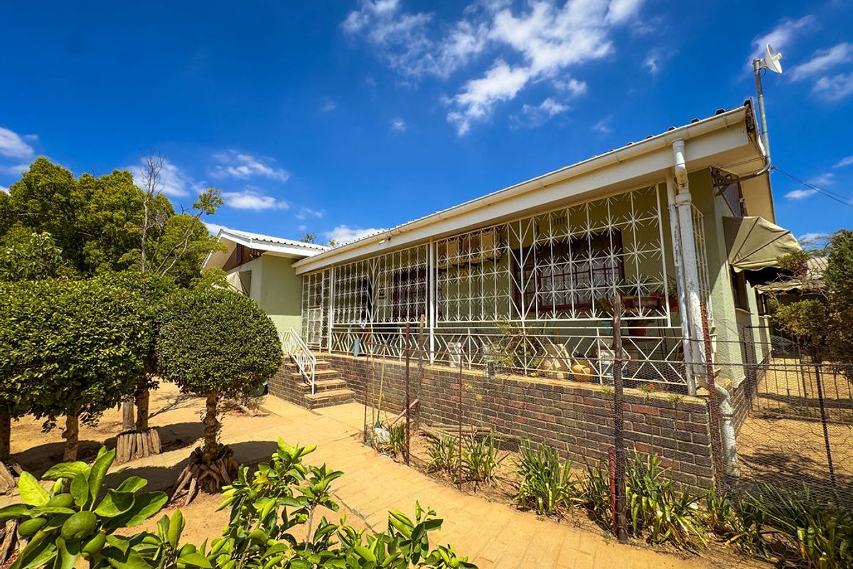 Rear view of the house featuring a covered patio, brick foundation, and security bars, set within a garden area with a paved walkway.
